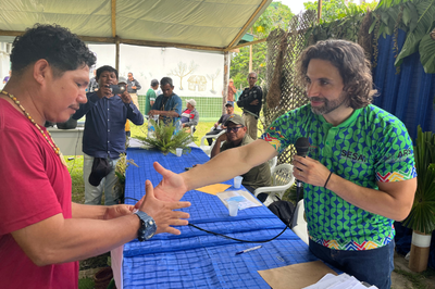 Homem com camisa verde estampada e microfone interagindo com um homem de camisa vermelha escura com um colar indígena em uma mesa coberta com tecido azul, em um evento.