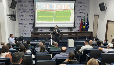 Momento da audiência pública. A imagem mostra uma sala de conferência formal, com várias pessoas sentadas e voltadas para um palestrante. O palestrante está em pé, usando microfone, diante de uma tela de projeção grande