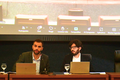 Os procuradores Lucas Dias (esq.) e Luidgi Merlo (dir.) debatem sobre a regulamentação do uso sacramental da ayahuasca durante audiência pública na sede do MPF em Rio Branco (AC), em 28/11/2025. Foto: Comunicação/MPF. A foto anexa mostra dois homens, sentados lado a lado em uma mesa de conferência, provavelmente durante uma audiência pública ou evento formal que ocorre online, como sugerido pela tela de projeção atrás deles. O homem à esquerda, com barba escura e vestindo um blazer verde-oliva sobre uma camisa clara, está falando, com a boca aberta em meio a uma frase. O homem à direita, usando óculos e com barba e cabelo escuros, veste um terno escuro sobre uma camisa branca e olha para frente ou ligeiramente para baixo, enquanto ambos têm laptops abertos à sua frente e copos de água em cálices ao lado. A tela de projeção no fundo exibe elementos de uma interface de reunião virtual, incluindo opções como "Participantes", "Chat", "Reagir", "Levantar mão", "Compartilhar", e "Mostrar legendas", e o nome do arquivo da imagem sugere que o evento é uma Audiência Pública sobre a regulamentação do uso sacramental da Ayahuasca, realizada pelo Ministério Público Federal (MPF) no Acre.