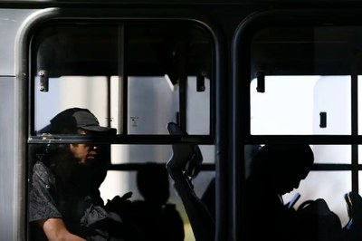 Fotografia das janela de um ônibus na qual pode-se ver passageiros sentados.