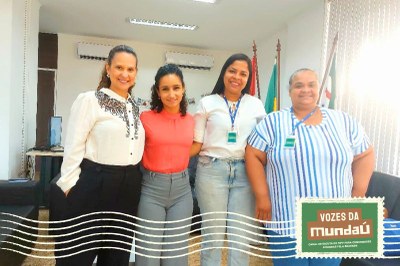 A foto mostra quatro mulheres posando lado a lado, sorrindo para a câmera em uma sala institucional. Todas usam crachás de identificação. Ao fundo, aparecem bandeiras, ar-condicionados e parte do mobiliário do ambiente.
No canto inferior direito, há um selo gráfico com o texto “Vozes da Mundaú".
