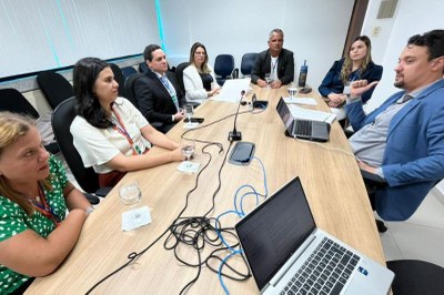 Na imagem, um grupo de sete pessoas está reunido em uma sala de reunião. Elas estão sentadas ao redor de uma mesa retangular de madeira clara. Todos parecem concentrados e engajados na conversa.

Há dois notebooks abertos, diversos cabos organizados no centro da mesa, copos com água e dois microfones de mesa, sugerindo uma reunião formal ou técnica.

As pessoas estão dispostas em ambos os lados da mesa, em um ambiente corporativo, com cadeiras pretas e uma parede branca ao fundo.