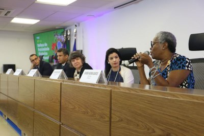 Foto: Comunicação/MPF imagem de mesa larga com cinco pessoas, em primeiro plano uma mulher negra de cabelos brancos fala ao microfone.