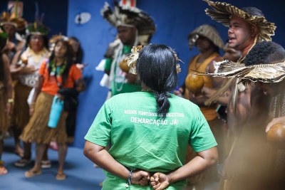 Antonio Augusto/MPF Uma imagem de um grupo de pessoas de uma tribo indígena, reunidas em uma sala.
A composição da cena mostra um grupo numeroso de pessoas indígenas, reunidas em uma sala. As pessoas estão posicionadas em um agrupamento, com algumas mais próximas ao centro da imagem e outras um pouco mais afastadas. Há uma diversidade de vestimentas e adereços, indicando possível pertencimento a uma cultura específica. As expressões faciais das pessoas presentes são variadas, mas a atmosfera geral parece ser de reunião ou celebração. O enquadramento é amplo, abrangendo grande parte do grupo, e a perspectiva é ligeiramente de baixo para cima, que cria uma sensação de respeito.
