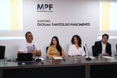 Imagem - Comunicação MPF Quatro pessoas estão sentadas lado a lado em uma mesa de auditório do MPF. À esquerda, um homem fala ao microfone diante de um laptop. Ao centro, duas mulheres estão sentadas, e à direita há um homem de terno. Ao fundo, aparece a parede com o nome “Auditório Gicelma Santos do Nascimento”.