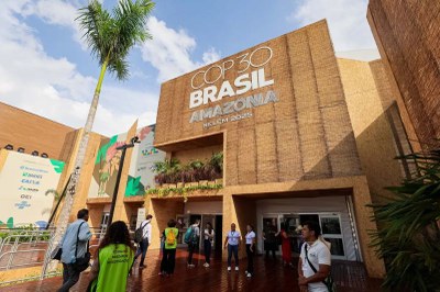 Fachada de um prédio coberto de madeira e bambu com os dizeres "COP30 Brasil Amazônia Belém 2025". Pessoas circulam na entrada.