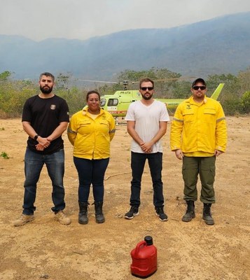 Foto de quatro pessoas em pé, com as mãos cruzadas na frente. Um homem e uma mulher vestem camisas amarelas. A mulher e um homem de camiseta branca estão no centro, com Marcelo à direita e um policial à esquerda. Ao fundo, um helicóptero amarelo claro, utilizado no combate às queimadas. Atrás do helicóptero, ve-se uma paisagem parcialmente queimada pelo fogo.