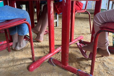 crianças sentadas em carteiras vermelhas em chão de terra batida 