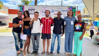 Um grupo de pessoas posa para foto em frente a um painel com logomarcas de entidades apoiadoras. O grupo se posiciona lado a lado, com braços dados ou próximos, aparentando união e descontração. O ambiente é externo e ensolarado, com tenda e estruturas de evento ao fundo.