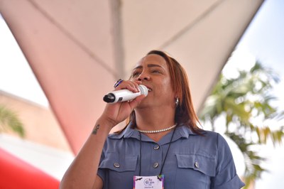 Uma pessoa segura um microfone e realiza uma fala em público, aparentando estar em um palco ou estrutura coberta. Ela veste camisa azul e acessórios como colar e brincos. A expressão facial transmite segurança e envolvimento com a atividade. Ao fundo, há elementos do ambiente externo, como árvores e o céu.