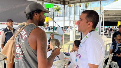Um homem vestindo camiseta sem mangas e boné conversa com outro homem de camiseta branca, aparentemente em um evento ao ar livre. Ambos parecem engajados em diálogo, com o segundo usando crachá e uniforme de serviço. Ao fundo, outras pessoas estão sentadas sob a cobertura, algumas atendendo ou aguardando atendimento.
