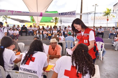 Uma equipe com coletes da Cruz Vermelha atende pessoas sentadas em mesas de triagem. Uma mulher com colete vermelho sorri enquanto manuseia documentos. No centro, um homem mais velho com chapéu aparenta aguardar informações. Ao fundo, um público numeroso está sentado assistindo ou aguardando atendimento. O clima é de ação social organizada.