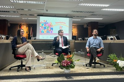 A imagem anexa captura um momento de um evento formal, possivelmente um seminário ou debate, que ocorre em um auditório ou sala de conferências. Três palestrantes estão sentados em cadeiras de escritório pretas sobre um pequeno palco elevado. No centro, um homem de meia-idade, vestindo um terno escuro com uma gravata vermelha, está falando ao microfone, segurando um papel. À sua esquerda (direita do observador), uma mulher de óculos, vestindo uma blusa azul-marinho e calças claras, está sentada com um telefone na mão. À direita dele (esquerda do observador), um homem mais jovem, com barba e vestindo uma camisa social azul clara e calças escuras, está sentado com as pernas cruzadas. Atrás deles, um projetor exibe um slide com o texto "SEMINÁRIO DIREITOS HUMANOS" e a frase "Por uma justiça plural e solidária", ilustrado com figuras coloridas representando diversidade. O palco é decorado com um arranjo floral de tons de rosa, vermelho e branco, e há copos de água e microfones de lapela para os participantes.