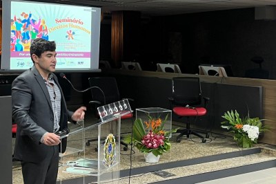 A imagem anexa captura um momento de um seminário ou evento formal sobre Direitos Humanos, conforme indicado pelo slide projetado ao fundo. O foco principal é um homem de pele clara, vestindo um terno escuro sobre uma camisa estampada, que está de pé atrás de uma tribuna de acrílico transparente e falando ao microfone. Ele gesticula com a mão direita, sugerindo que está proferindo um discurso ou palestra. No slide projetado, lê-se "Seminário Direitos Humanos" e o tema parece ser "Por um... antidiscriminatória", embora o texto esteja parcialmente ilegível. A sala é escura, destacando o palestrante e o slide, e apresenta uma disposição formal de assentos em fileiras vazias (algumas cadeiras pretas e uma vermelha), típicas de auditórios ou salas de tribunal/plenário. A tribuna é decorada com o que parece ser um brasão ou logo institucional, e há arranjos de flores tropicais em vasos adornando o púlpito e o palco elevado.