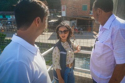 Três pessoas conversam em uma passarela ou área próxima ao canal. Ao centro, a mesma mulher de colete institucional e óculos escuros gesticula com a mão, aparentemente explicando algo. Dois homens a escutam atentamente, um à esquerda e outro à direita. Ao fundo, vê-se o canal, casas simples e moradores circulando na região.