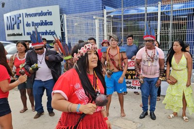 A imagem registra uma manifestação de lideranças indígenas em frente à sede da Procuradoria da República no Maranhão (MPF), em São Luís. No centro do registro, um grupo de homens e mulheres das etnias Guajajara e Timbira aparece trajando vestimentas tradicionais, como cocares de penas coloridas, pinturas corporais e colares, mesclados a roupas contemporâneas. Alguns seguram maracás, instrumentos rituais utilizados durante o ato. Ao fundo, é possível ver a fachada do prédio público com identificação oficial e outros manifestantes segurando uma faixa, sugerindo uma mobilização por direitos ou demandas territoriais junto ao Ministério Público Federal.