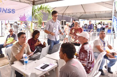 Um grupo de pessoas está sentado e em atendimento em uma mesa, com notebooks à frente. Uma pessoa em pé, segurando cabos, parece auxiliar no processo. Há interação entre os participantes e atendimento ao público. O ambiente é uma estrutura coberta, com fluxo de pessoas e clima de mutirão de serviços.