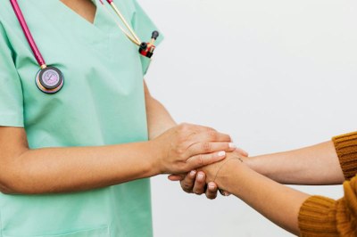 Foto ilustrativa: Canva Foto em detalhe de uma profissional da saúde com roupa verde de hospital e estetoscópio no pescoço segurando as mãos de uma paciente usando uma roupa amarela mostarda