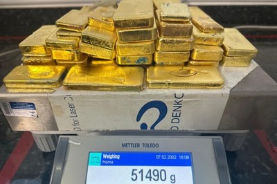A imagem anexa exibe uma pilha de barras de ouro polidas e retangulares, empilhadas de forma um tanto desordenada, sobre uma balança digital de precisão da marca Mettler Toledo. A tela da balança, que mostra a data 07.02.2002 e a hora 18:08, indica um peso total de 51490 g, o que equivale a mais de 51 quilos. O ambiente onde a pesagem ocorre parece ser um laboratório ou um local de custódia, sugerindo uma transação ou uma apreensão de alto valor. A iluminação destaca a cor dourada intensa e os reflexos do metal precioso.