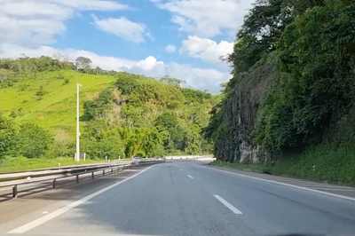  fotografia mostra um trecho da rodovia BR-040 no Rio de Janeiro. A cena é dominada por uma paisagem natural exuberante, com a estrada asfaltada curvando-se suavemente pelo centro. À esquerda, uma encosta gramada e verdejante sobe em direção ao topo, com uma palmeira de luz visível. No lado direito da estrada, um paredão rochoso abrupto e coberto por vegetação densa delimita o caminho. O céu é claro, com algumas nuvens brancas, sugerindo um dia ensolarado, e a vegetação tropical é farta e em tons variados de verde, enfatizando o ambiente serrano ou montanhoso da região.

