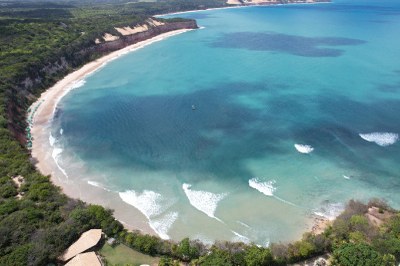 Foto das falésias de Pipa, tomada de cima, de um drone, a partir do continente, de onde se vê a vegetação por sobre as falésias, bem como parte da praia e o mar em frente, formando uma curva côncava na praia"