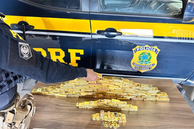 Foto: Polícia Rodoviária Federal Foto mostra dezenas de barras de ouro empilhadas sobre uma mesa em frente a uma viatura da Polícia Rodoviária Federal (PRF), com um policial fardado ao lado.