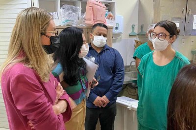 Foto: Comunicação/MPF Pessoas com máscaras, jalecos verdes e roupas casuais conversam em uma sala com armários e materiais médicos ao fundo.