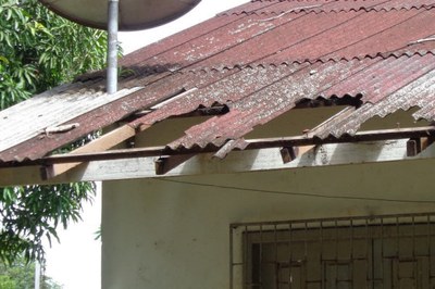 Telhado de uma das escolas (foto: Funai) foto de detalhe de um telhado de amianto vermelho desgastado e quebrado, com partes faltando, sobre uma parede branca e uma janela gradeada.