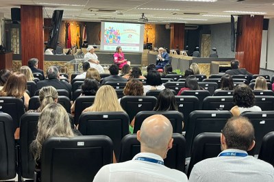 A procuradora da República Janaina Andrade representa o MPF no Seminário de Direitos Humanos – Por uma Justiça Plural e Antidiscriminatória, no auditório do TRT-PB, em João Pessoa (PB), em 5/12/2025. Foto: Comunicação/MPF. Auditório com paredes de madeira, cadeiras pretas ocupadas por público atento. No palco, três pessoas debatem diante de uma tela que exibe o título "Seminário Direitos Humanos: Por uma justiça plural e antirracista". Há bandeiras e um crucifixo ao fundo, indicando ambiente institucional.