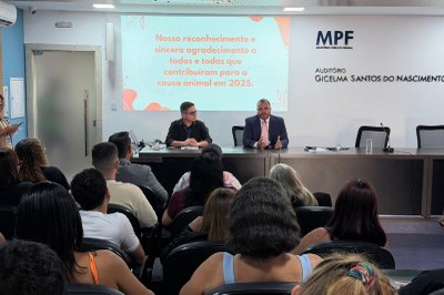 Imagem do auditório do evento mostra público e na frente a mesa, com um telão atrás