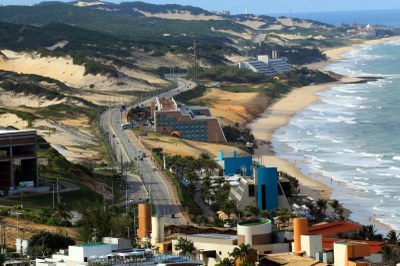Imagem feita do alto, registrando parte da Via Costeira, com sua pista, hotéis e praias, tendo o Parque das Dunas à esquerda e o mar à direita, do ponto de vista de quem se encontra em Ponta Negra.
