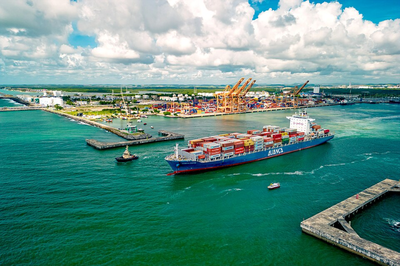Foto aérea do Complexo Portuário de Suape, mostrando um grande navio de contêineres sendo auxiliado por um rebocador em águas verde-esmeralda, com o terminal e guindastes ao fundo.