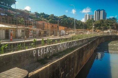 Vista externa de um canal urbano com água correndo em seu leito. À margem esquerda, há um muro de contenção com uma grade metálica acima, separando o canal de uma área com construções residenciais. No muro, é possível ler a frase “PEKENO – VAMOS CUIDAR” escrita em tinta branca. Ao fundo, aparecem casas em encosta e edifícios mais altos, compondo uma paisagem urbana.