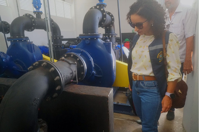 Foto: Comunicação MPF Uma mulher usa colete institucional e óculos escuros enquanto observa um conjunto de grandes tubulações e válvulas industriais de cor azul, instaladas em um ambiente fechado. Ela está de pé ao lado dos equipamentos, olhando atentamente para as estruturas metálicas, que aparentam fazer parte de um sistema hidráulico ou de bombeamento. Ao fundo, outra pessoa acompanha a visita técnica.