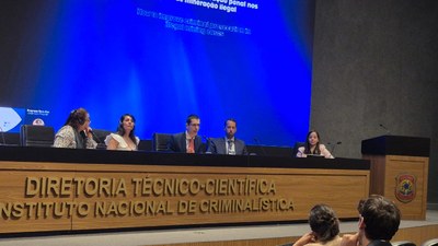 Mesa de debates em um auditório do Instituto Nacional de Criminalística. Seis pessoas estão sentadas atrás de uma bancada, participando de um painel. Ao fundo, um telão azul exibe o tema sobre aprimoramento da persecução penal em casos de mineração ilegal, enquanto o público acompanha a apresentação.