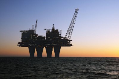 Foto ilustrativa: morkeman/Getty Images/Canva Silhueta de uma plataforma de petróleo iluminada sobre o mar escuro, sob céu azul que se torna alaranjado no horizonte.