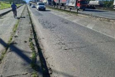 Foto mostra trecho de uma via marginal de uma rodovia. O asfalto está rachado e esburacado. 