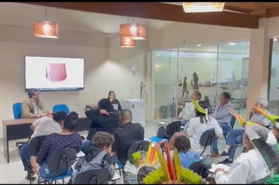 MPF se reúne com indígenas e representantes da prefeitura de Armação dos Búzios (RJ), na sede da Secretaria Municipal de Turismo, em 27/11/2025. Foto: Comunicação/MPF. A imagem anexa mostra uma reunião ou apresentação em ambiente interno com aproximadamente 15 a 20 pessoas. O foco da sala parece estar em uma tela de televisão/monitor à esquerda, que exibe uma imagem em close-up de um objeto com textura de cestaria ou tecido. À frente da tela, um homem de blazer e óculos está sentado a uma mesa, conversando com uma mulher que está de pé ou sentada na mesma mesa. A maioria dos participantes está sentada em cadeiras dispostas em filas, e alguns deles, em primeiro plano à direita, usam adornos de cabeça indígenas com penas coloridas, indicando a participação de membros de povos originários no evento. O ambiente é bem iluminado, tem um aspecto moderno e, ao fundo, há uma parede de vidro que permite ver uma área externa ou um diorama com figuras que parecem representar pessoas.