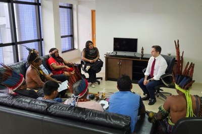 O procurador-chefe do MPF no Maranhão, Alexandre Soares (centro), realiza reunião com indígenas Guajajaras e Timbiras, na sede do órgão, em São Luís (MA), em 19/12/2025. Foto: Comunicação/MPF. A imagem registra uma reunião institucional em um escritório, na sede do Ministério Público Federal no Maranhão, em São Luís. No centro da cena, diversas lideranças indígenas dos povos Guajajara e Timbira, vestindo trajes tradicionais e cocares de penas coloridas, estão sentadas em um sofá preto em formato de "L" e em cadeiras de escritório. Eles dialogam com o procurador-chefe do MPF no Maranhão, Alexandre Soares, em vestes formais (camisa branca e gravata vermelha) e uma mulher posicionada ao centro, que parece mediar a conversa. O ambiente é sóbrio, com janelas amplas ao fundo, um televisor desligado sobre um balcão de madeira e uma atmosfera de negociação ou audiência oficial voltada para questões das Terras Indígenas Bananal e Bacurizinho.