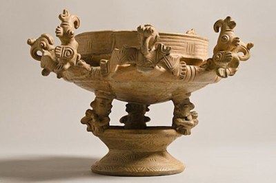 Vaso de cerâmica bege sobre fundo neutro. Três figuras de base sustentam a tigela, decorada com relevos e esculturas de pássaros na borda.