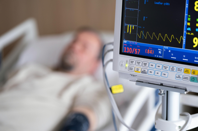Foto de ambiente hospitalar mostra, em primeiro plano, um monitor de sinais vitais e, em segundo plano desfocado, um homem deitado em cama hospitalar