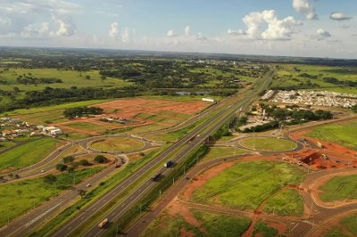 Trecho de duplicação da BR-153 (Transbrasiliana) em São José do Rio Preto (SP), em fevereiro de 2022. Foto: Dnit. A imagem anexa apresenta uma vista aérea de um trecho da Rodovia BR-153 em São José do Rio Preto, São Paulo, destacando as obras de duplicação e infraestrutura viária em andamento, conforme sugerido pelo nome do arquivo. No centro, a rodovia duplicada se estende pelo cenário, com tráfego de veículos, incluindo caminhões. A via é ladeada por grandes áreas de terraplanagem e desenvolvimento imobiliário (lotes de terra avermelhada), indicando crescimento urbano na região. O design da estrada é complementado por interseções e rotundas (rotatórias) bem definidas, que buscam organizar o fluxo de veículos de e para a rodovia. O ambiente é predominantemente verde, com extensas áreas de pastagem ou vegetação nativa ao redor da área de construção. O céu é parcialmente azul, pontilhado por nuvens brancas e fofas, sugerindo um dia claro e ensolarado.