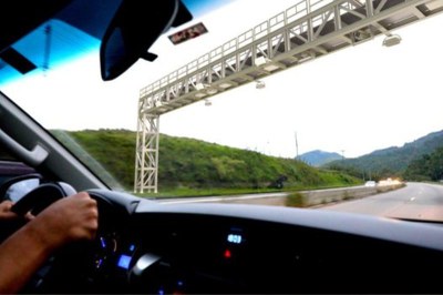 Foto mostra em detalhe motorista dentro de um carro passando por baixo de um sistema free flow na rodovia