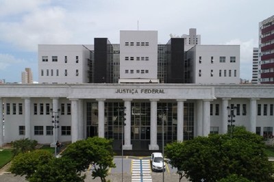 Foto: JFRN Foto da fachada da Justiça Federal do Rio Grande do Norte, tomada de frente e do alto, mostrando o prédio em cor predominantemente branca, com grandes pilares, o nome Justiça Federal na parte superior e aparecendo ainda parte dos jardins