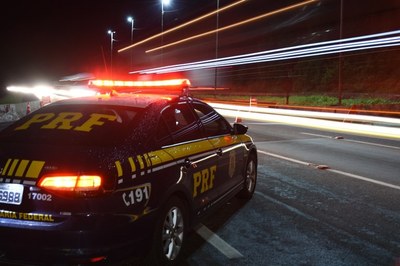 Viatura da Polícia Rodoviária Federal (PRF). Foto: PRF. Esta imagem captura uma viatura da Polícia Rodoviária Federal (PRF) estacionada à beira de uma rodovia durante a noite, com seu giroflex acionado emitindo uma luz avermelhada intensa que ilumina a traseira do veículo. O automóvel, de cor escura com os tradicionais grafismos amarelos da corporação e o número de emergência 191 visível na lateral, está posicionado em primeiro plano, enquanto o fundo é composto por longos rastros de luz branca e alaranjada deixados pelo tráfego em movimento, técnica característica de fotografias com longa exposição. A cena, composta sob a iluminação dos postes da estrada, transmite uma sensação de vigilância e segurança viária em meio à escuridão.