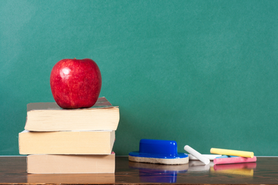 Foto ilustrativa: Canva Foto de uma maçã vermelha no topo de uma pilha de três livros, com um apagador azul e giz sobre a mesa, em frente a um quadro-negro verde.
