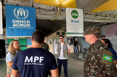 Integrante do MPF e militar do Exército conversam com profissionais de ajuda humanitária em um abrigo, com banners do ACNUR e da AVSI Brasil ao fundo.