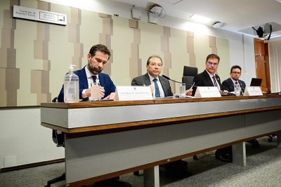 Foto mostra quatro homens sentados atrás de bancada de auditório