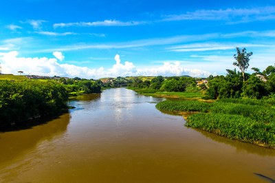Rio Paraíba do Sul. Foto: Claudio Vieira / Pref. São José dos Campos #PraTodosVerem: vista panorâmica do rio Paraíba do Sul sob um céu azul vibrante com nuvens brancas; as águas calmas e acastanhadas são ladeadas por densa vegetação verde em ambas as margens, com colinas suaves e construções urbanas ao fundo.