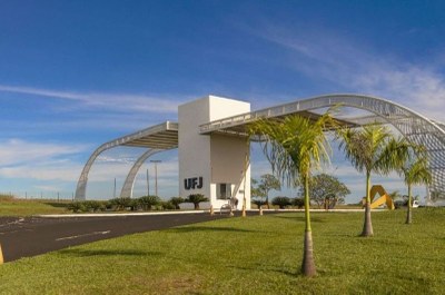#PraTodosVerem: fotografia da entrada da Universidade Federal de Jataí. Em primeiro plano, aparecem palmeiras pequenas em um gramado. O elemento central é um portal moderno de estruturas brancas curvas e uma torre com a sigla "UFJ", que atravessa a via principal de acesso.